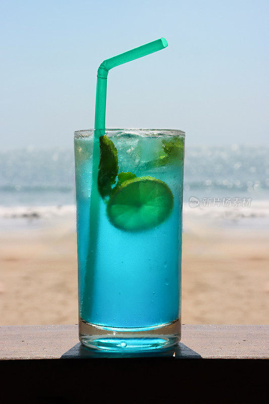 在露天海滩酒吧餐厅的木栏杆上的鸡尾酒特写图像，蓝色泻湖伏特加和蓝色cura<s:1>鸡尾酒，柠檬片，薄荷叶，吸管，沙子和大海的背景